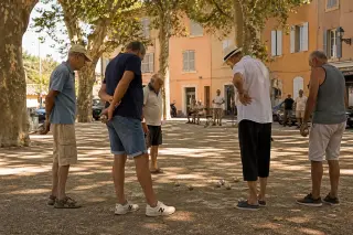 Group of pétanque players chatting around a cochonnet under shady plane trees on a Provençal village square with warm Mediterranean light.
