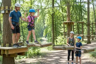 Parcours d’accrobranche en forêt : une enfant casquée traverse un pont de rondins, aidée par un adulte, sous le regard d’une mère et de son fils