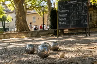 Close-up of shiny pétanque balls on a gravel boulodrome under dappled afternoon light, with a handwritten scoreboard and plane trees in the background.